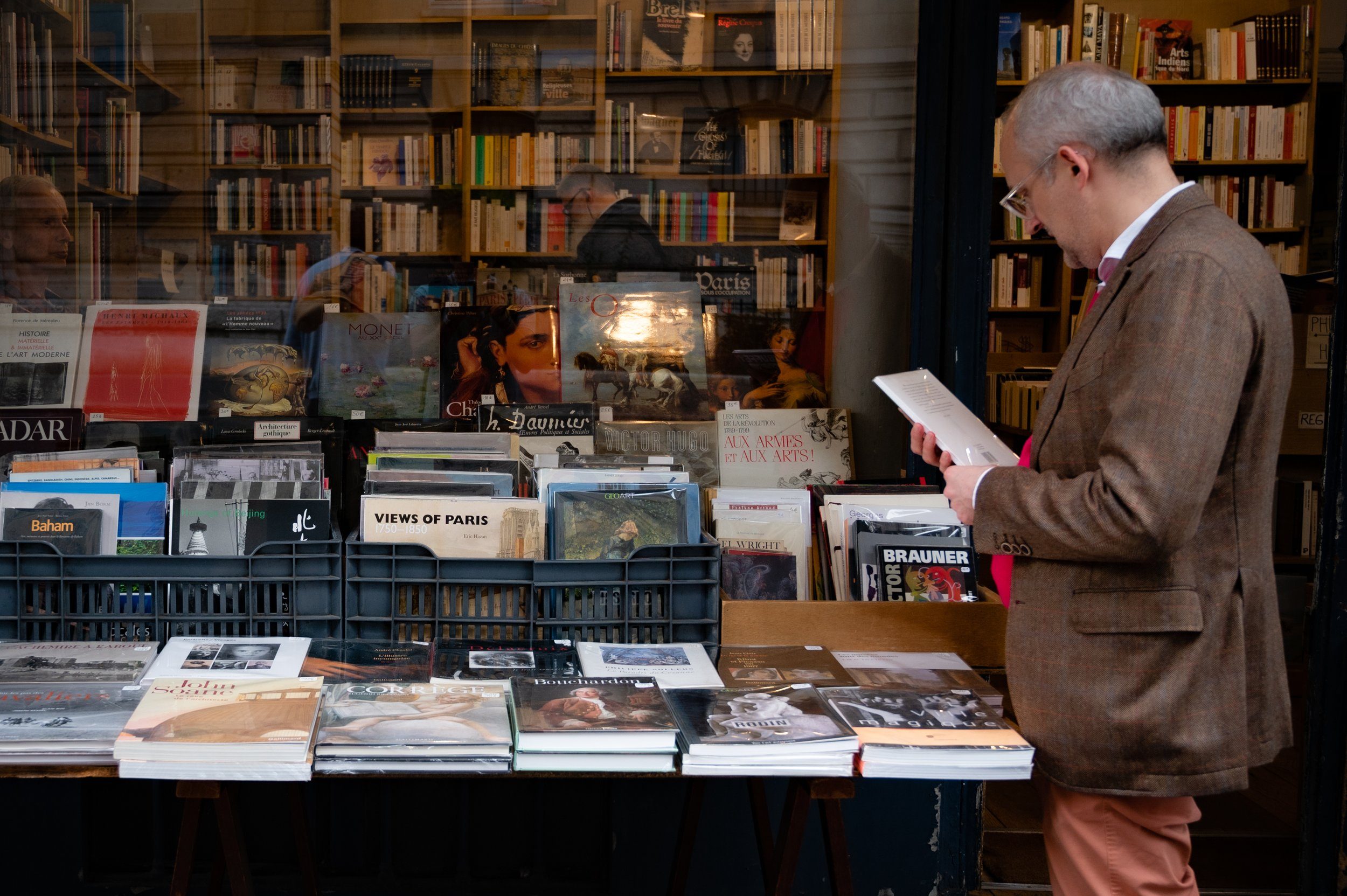 A man reading at a bookshop window in Paris, tables of secondhand books and prints on the pavement in front of him