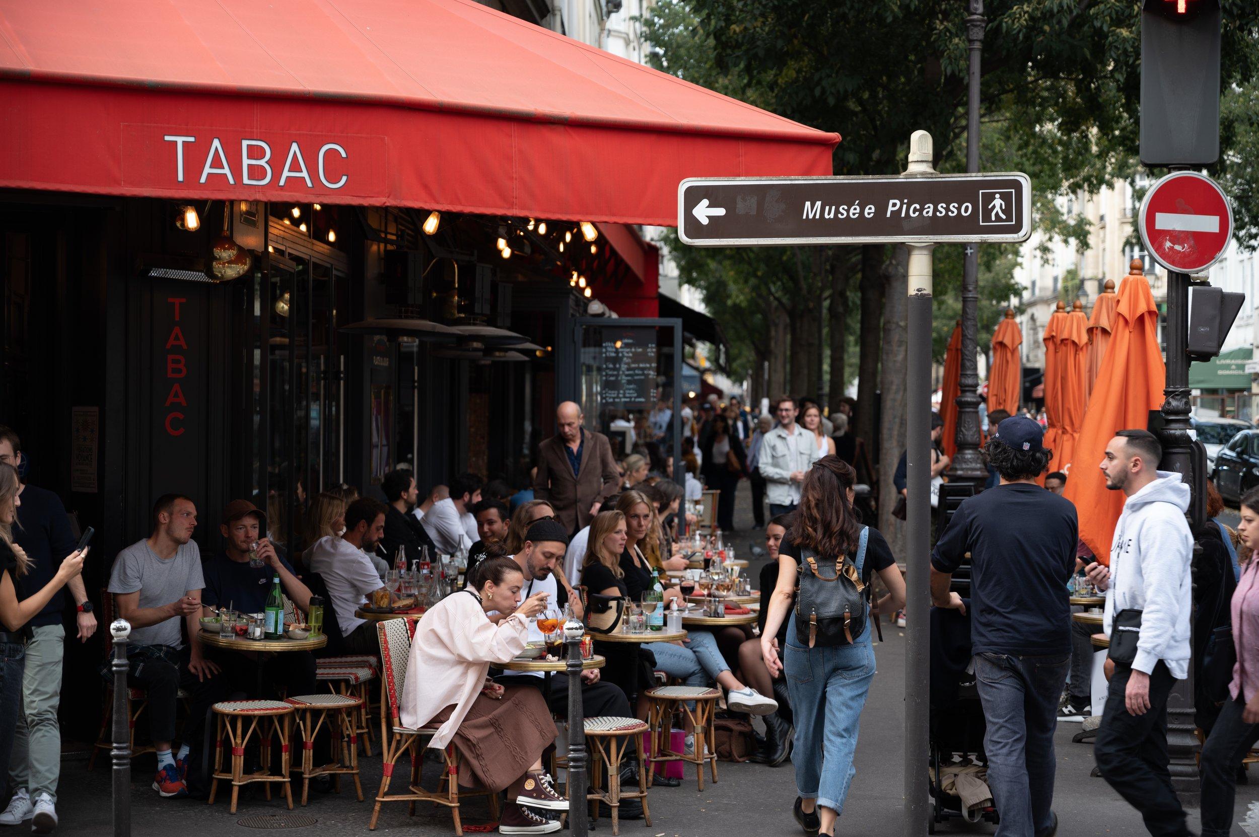 A packed pavement terrace under a red tabac awning in the Marais, a Musée Picasso direction sign on the pole beside it