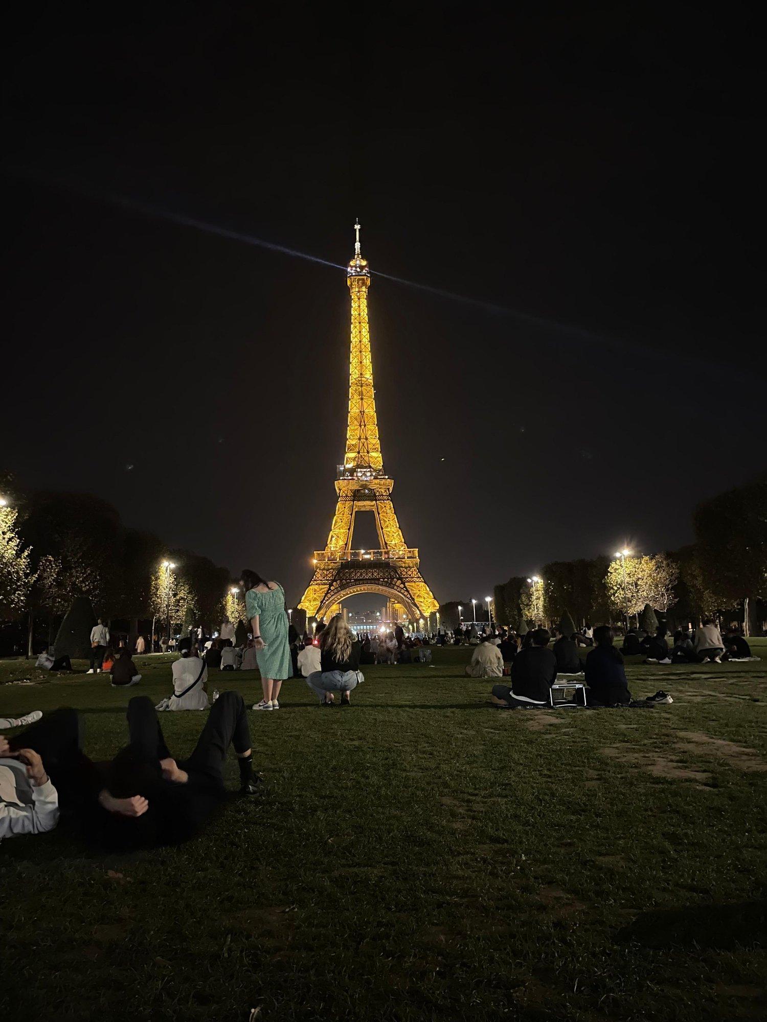 The Champ de Mars at night, the Eiffel Tower lit gold at the far end and people sitting out on the grass