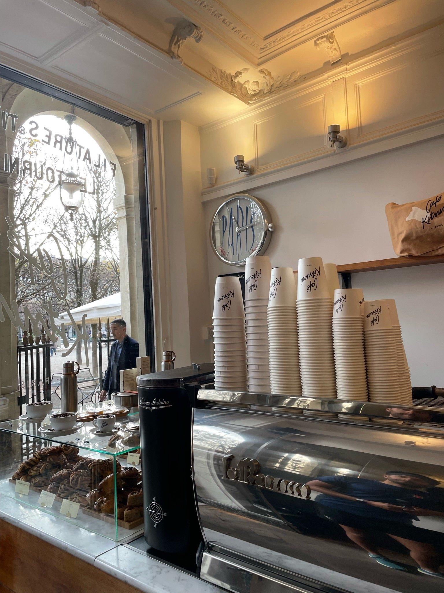 The counter inside Café Kitsuné, stacked branded cups beside the espresso machine and pastries under glass