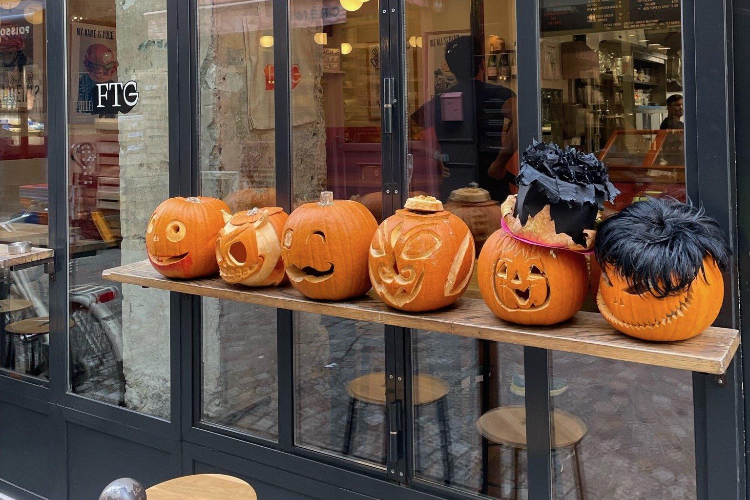 Carved pumpkins along the window ledge of Frenchie To Go on Rue du Nil, stools and the counter visible through the glass