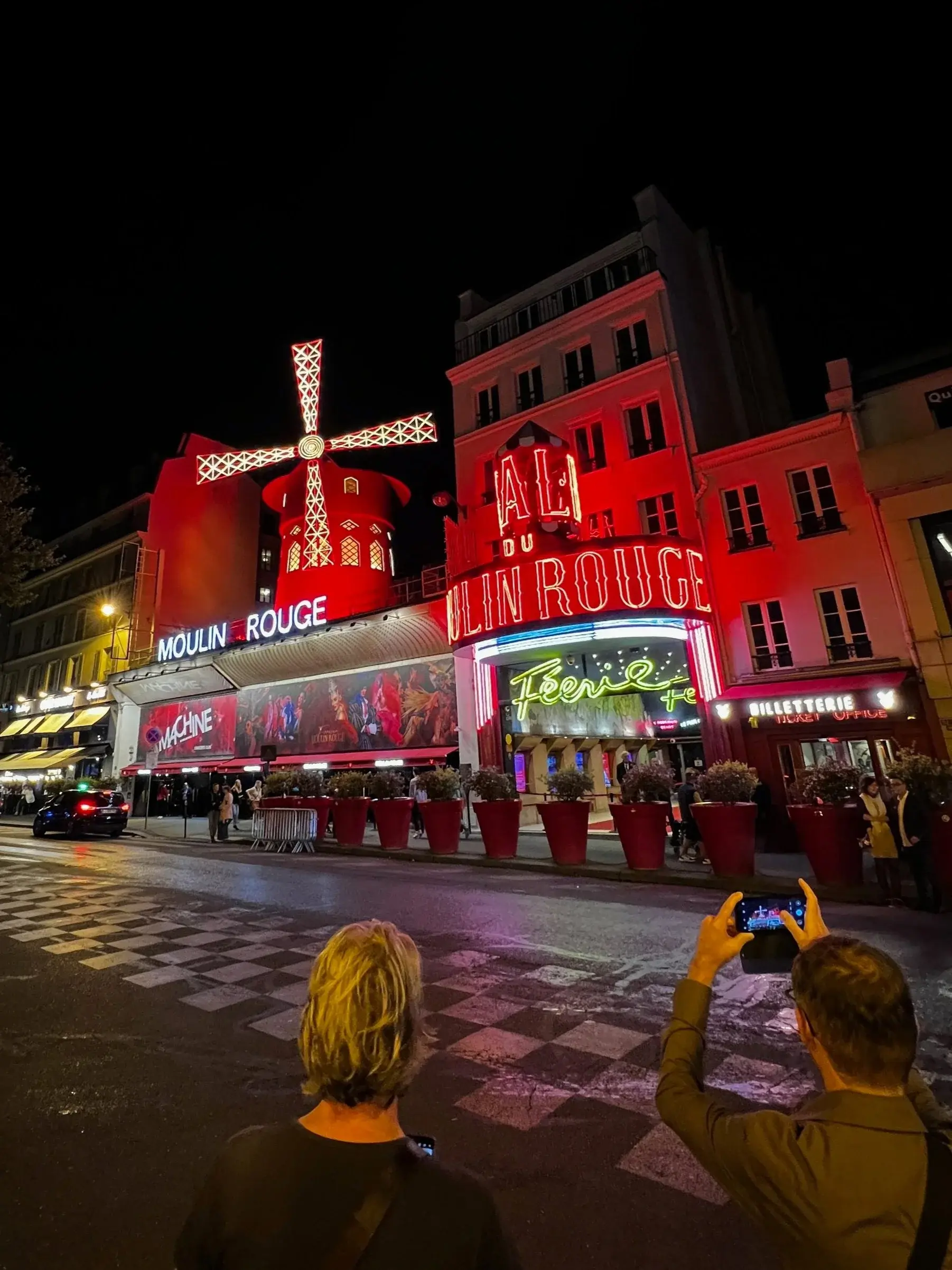 The Moulin Rouge's red windmill and neon frontage lit at night, people photographing it from across the boulevard