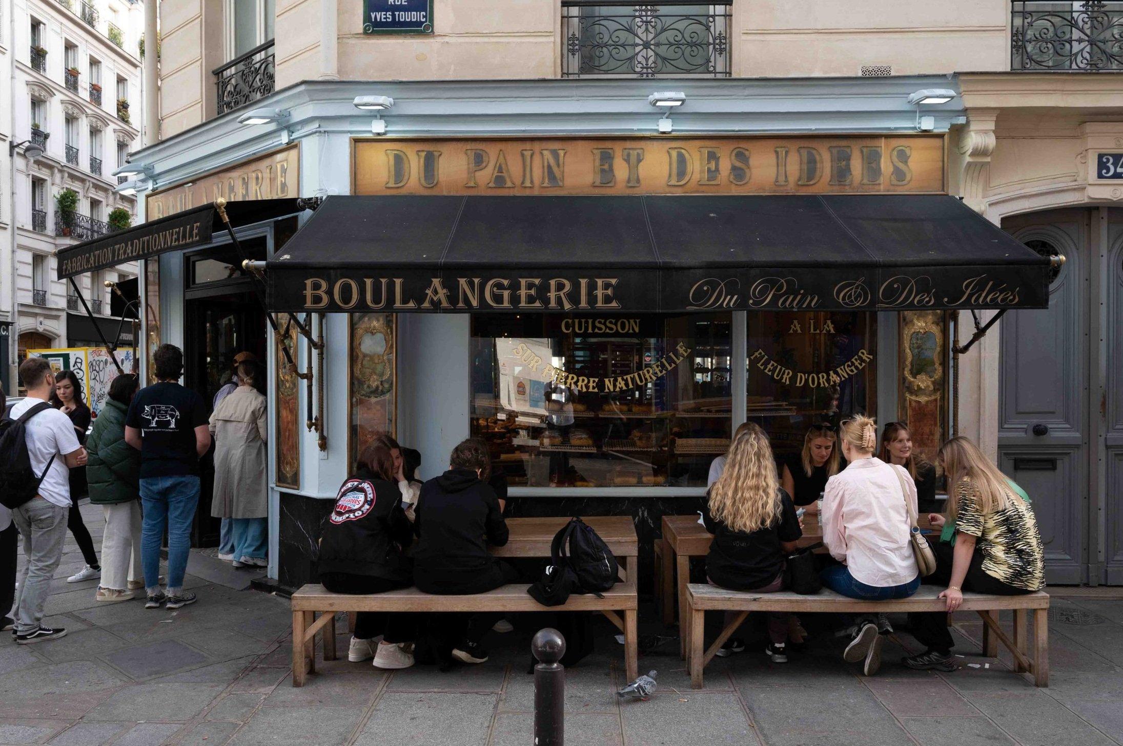 The gold-lettered corner shopfront of Du Pain et des Idées, people queuing at the door and sitting on the benches outside